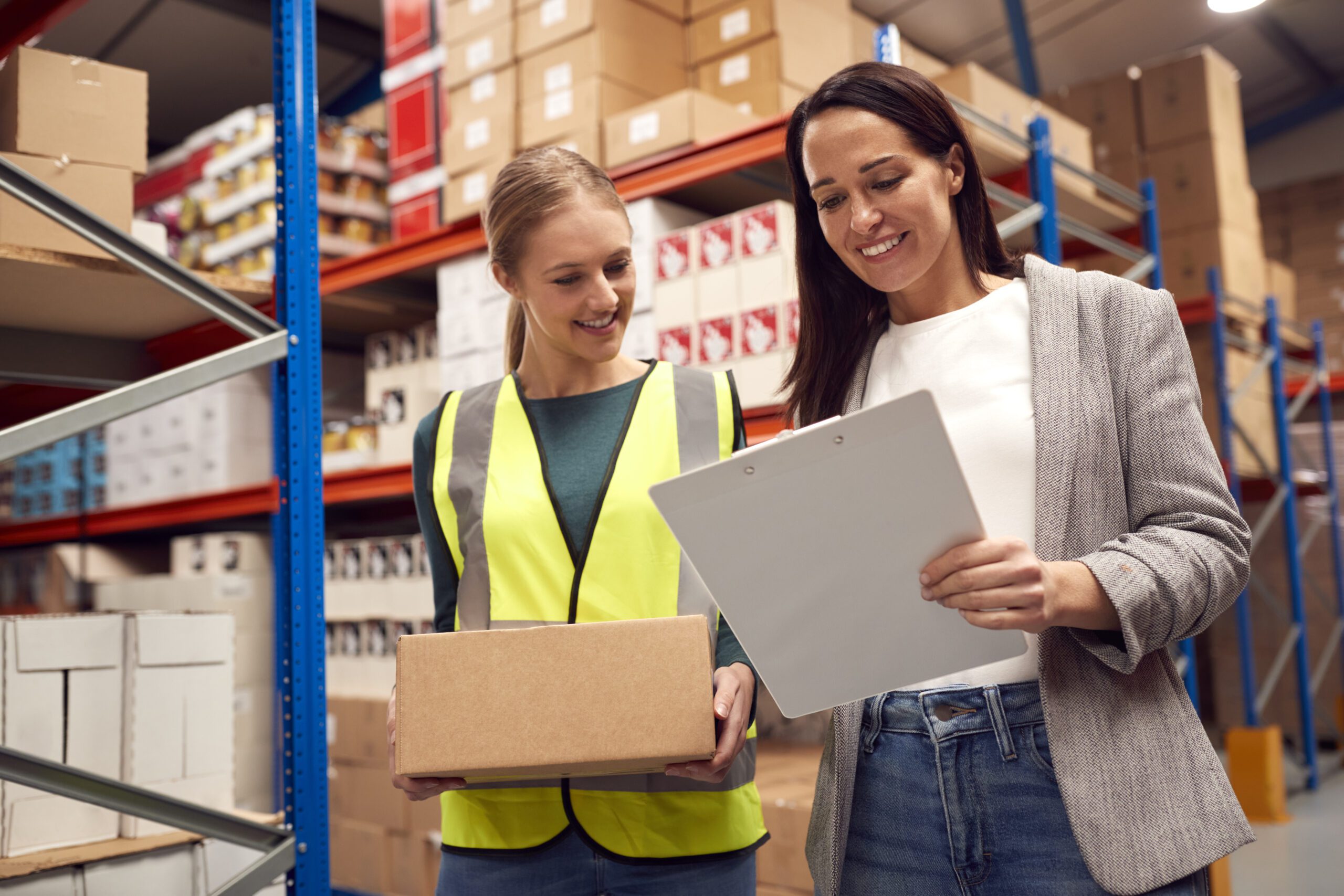 Female Team Leader With Clipboard In Warehouse Training Intern Standing By Shelves