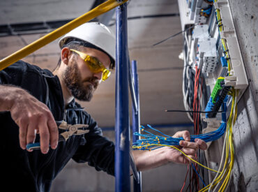 A male electrician works in a switchboard with an electrical connecting cable, connects the equipment with tools.