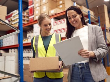 Female Team Leader With Clipboard In Warehouse Training Intern Standing By Shelves