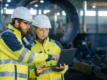 Male and Female Industrial Engineers in Hard Hats Discuss New Project while Using Laptop. They Make Showing Gestures.They Work in a Heavy Industry Manufacturing Factory.